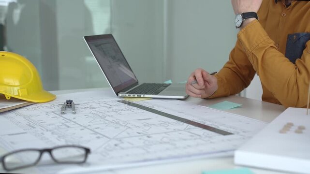 Engineer working on architectural blueprints and plans, typing on a laptop with a compass, ruler, safety helmet, glasses, and a building model on the desk