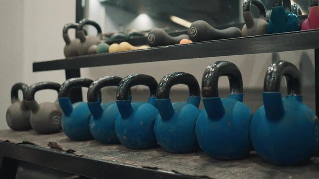Low angle close up shot of blue and black kettlebells neatly organized on a storage rack inside a commercial gym facility.