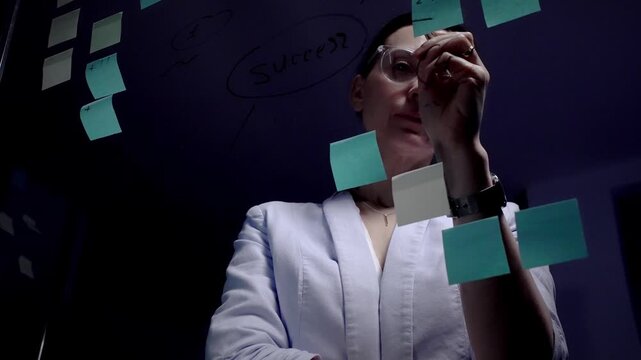 Woman wearing clear frame glasses standing in front of a glass wall covered with sticky notes and drawing a mind map, concentrating on planning a business strategy