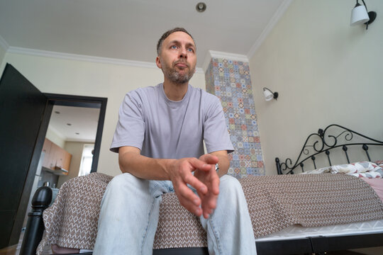 Worried man sits on bed at home looking into distance while thinking about uncertain future, personal problems and important decision.