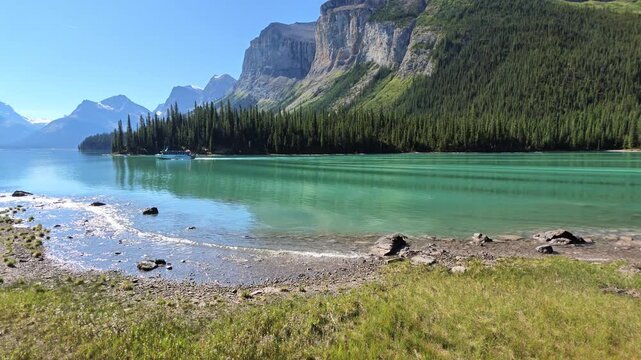 Spirit Island jutting into turquoise Maligne Lake with tour boat, surrounded by evergreen forest and snow-capped Canadian Rockies under clear blue summer sky in Jasper National Park, Alberta of Canada