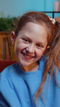 Close-up portrait of happy smiling Caucasian preteen school girl 10 years old. Young lovely child kid looking at camera at home night play room apartment sitting on orange couch. Ponytail hairstyle