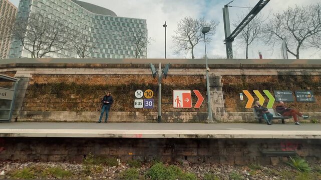 Scenic view from a moving train window passing urban graffiti walls, modern skyscraper towers, and waiting commuters along the platform of Javel station in Paris, France, during daytime.

