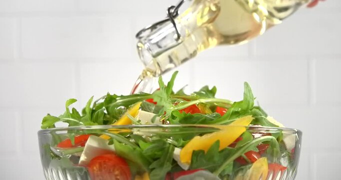 Sunflower oil pouring from bottle onto fresh vegetable salad with arugula tomato pepper and white cheese