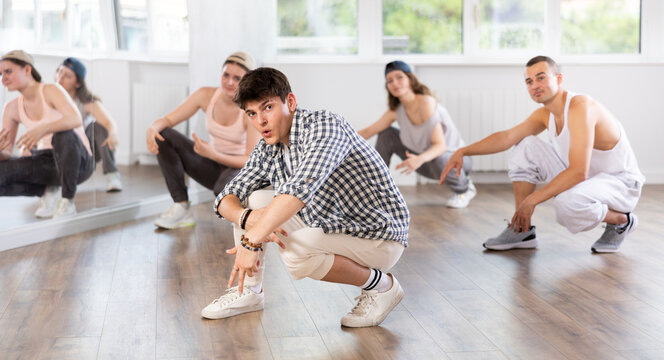 Positive young man engaged in break-dance squatting down together with other attendees of dancing courses