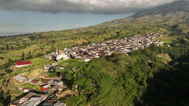 Aerial view of Kruwisan Village nestled in the lush mountainous landscape with terraced fields and a mosque featuring a golden dome in Kledung, Central Java, Indonesia.
