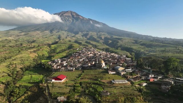 Aerial view of Kruwisan Village nestled at the foot of Mount Sumbing with lush terraced fields under a clear blue sky in Kledung, Central Java, Indonesia.