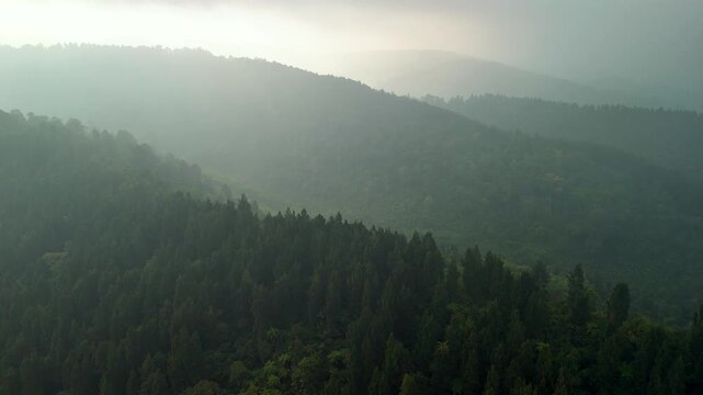 Aerial view of a lush, forested mountain range shrouded in mist and fog under a soft, overcast sky in Sukabumi, West Java, Indonesia.