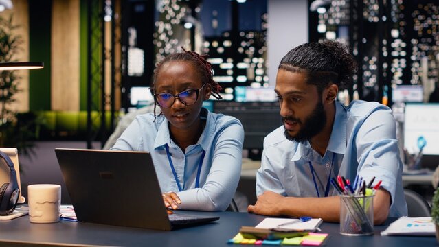 Diverse manager and assistant read inbox emails on a laptop late at night, analyze data and statistical information, and complete accounting assignments in a corporate office after normal hours.