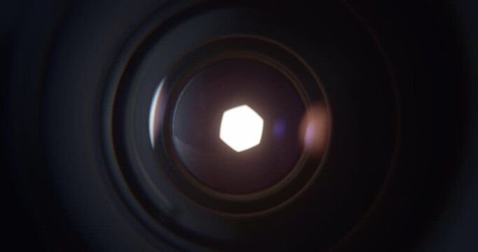 Extreme closeup of a camera lens aperture slowly opening as intense bright light emerges from behind the blades. Cinematic visual representing photography, optics, focus, exposure, illumination.
