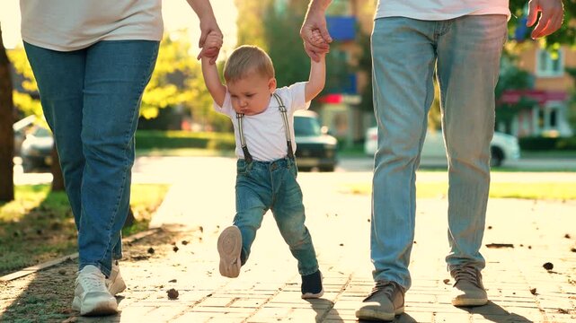Parents, child walk hand in hand, Baby first steps, city park. Mom dad kid son playing outside. Happy family. Dad mom child boy walking in park, summer. Family weekend. Baby son learns walk, sun day