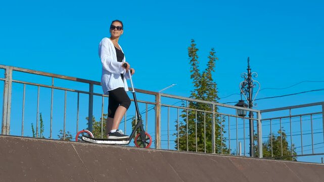 Modern woman with scooter. Woman in sportswear poses on a bridge with her scooter, soaking up the summer sunshine and enjoying the clear blue sky, embodying an active and healthy lifestyle