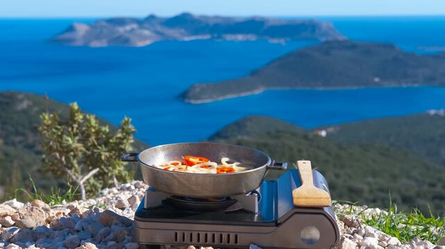 Outdoor cooking on portable gas stove overlooking the sea. Vegetables sizzling in a pan on a portable camping stove with a stunning aerial view of the blue sea and green islands