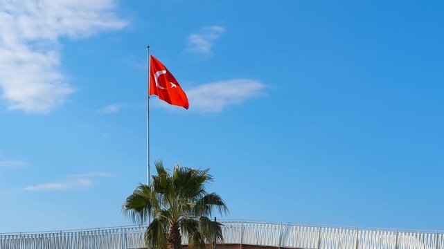 Turkish flag waving on a flagpole. Vibrant red turkish flag featuring white star and crescent, gently waving on flagpole against cloudless blue sky, framed by small palm tree and white fence