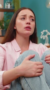 Young woman sits on home sofa hugging legs close feeling depression and stress after breakup job loss and bankruptcy. Caucasian girl stares downward apathetic on bad day seeking comfort in silence