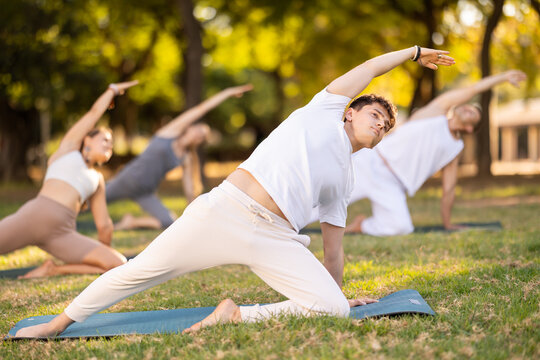 In serene setting of summer park, focused Hispanic guy practicing Reverse Gate Pose during group session, kneeling on one leg with arm reaching overhead, stretching side body and improving flexibility