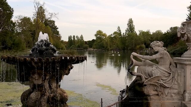 Static scenic view of an ornate fountain beside a classical female stone statue in the Italian Gardens, Hyde Park, London, with the park lake and greenery in the background.