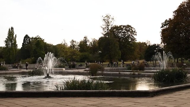 Wide scenic view of the Italian Gardens in Hyde Park, London, featuring ornamental fountains and landscaped surroundings as a pigeon flies across the frame in slow motion.