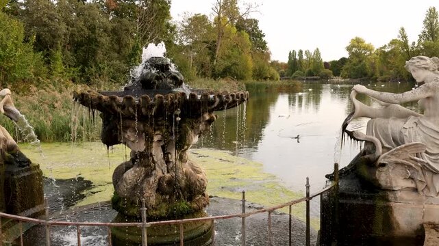 Slow panning shot across an ornate fountain and classical stone statues in the Italian Gardens, Hyde Park, London, with the park lake and autumn greenery in the background.