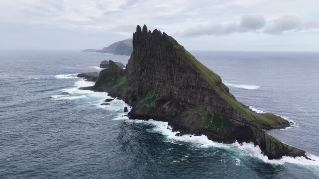 Aerial view of the dramatic Tindholmur sea stack rising from the North Atlantic Ocean with steep grassy cliffs and crashing waves in the Faroe Islands.