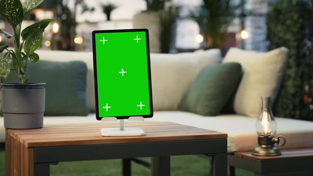 Mockup tablet with isolated screen beside a potted plant on a wooden rooftop terrace table, showing a close-up chroma key device in a high rise urban lounge in front of a cushioned couch.
