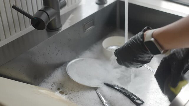 Woman wearing black gloves scrubs spoon with sponge and rinses it under running water in soapy sink. Cleaning cutlery during everyday dishwashing