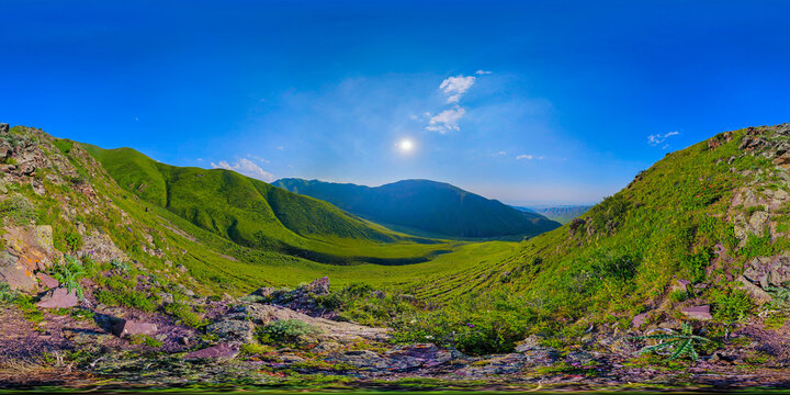 Full spherical 360-degree panorama captures a mountainous landscape in Kyrgyzstan. Features rocky terrain in foreground, rolling green hills, and a clear blue sky with sunlight.