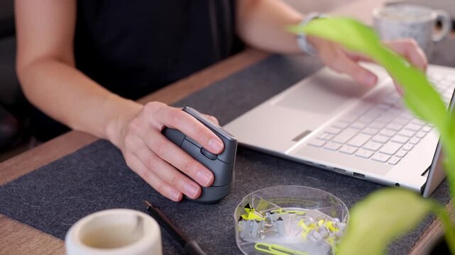 Woman using vertical ergonomic mouse and typing on laptop at desk. Closeup of hands switching between mouse and keyboard. Remote work and home office
