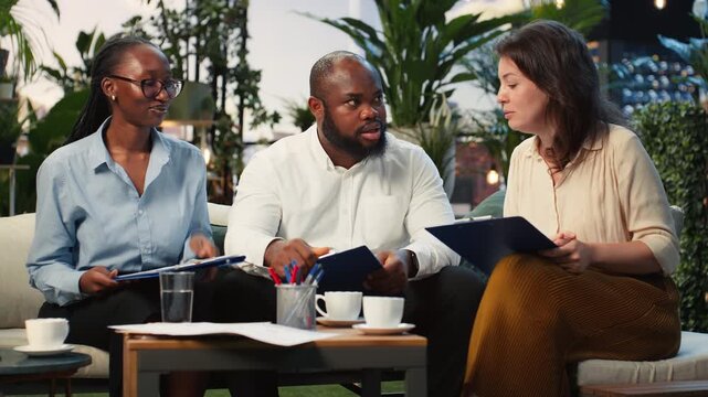 Smiling coworkers discuss project documents during a rooftop terrace meeting, comparing information as a business team against a downtown skyline.