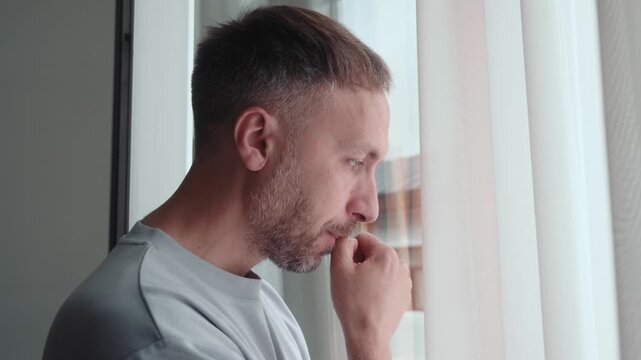 Worried man holds fingers near mouth while staring through window, feeling anxious and nervous as stress, uncertainty and emotional tension build at home.