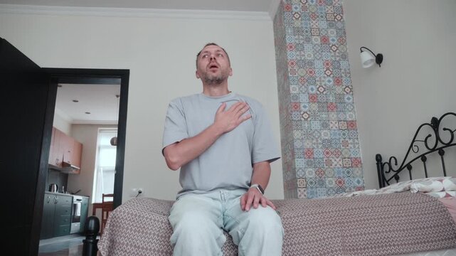 Man experiences panic attack or sudden chest discomfort at home, holding hand over heart and breathing heavily with shortness of breath and visible distress.