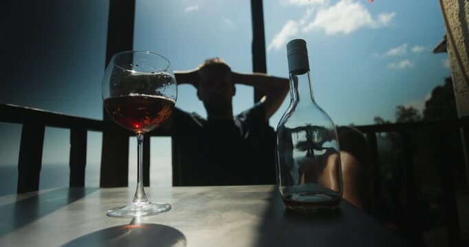 A glass of red wine and an empty bottle sit on an outdoor table while a person in silhouette relaxes against a bright blue sky, enjoying a moment of peace and contemplation.
