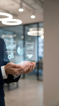 Businessman holding holographic display with growth charts, sustainability leaf icon, gears, and handshake symbols in modern office with ambient lighting