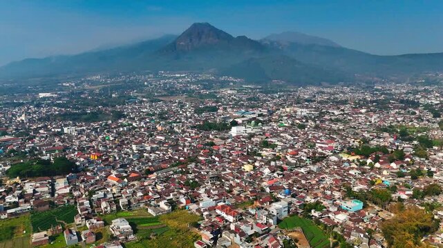 Aerial view of Batu city with the majestic Mount Panderman in the background, featuring dense urban rooftops and surrounding green landscape in Batu, East Java, Indonesia.