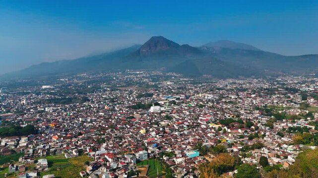 Aerial view of the sprawling city of Batu nestled at the foot of the majestic Mount Panderman, featuring dense urban rooftops and lush green surroundings in Batu, East Java, Indonesia.