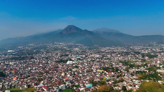 Aerial view of the urban landscape of Batu city nestled at the foot of the majestic Mount Panderman under a clear blue sky in Batu, East Java, Indonesia.