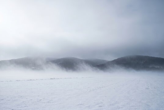Soft winter fog rolling across snow covered meadow with distant low hill silhouette barely visible in mist. Minimalist cold landscape with diffused light and empty copy space for seasonal background