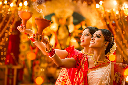 Devotional Dance of Lights: Two women adorned in traditional attire perform an Aarti ceremony, holding clay lamps aloft amidst the vibrant lights and decorations of a festive celebration.