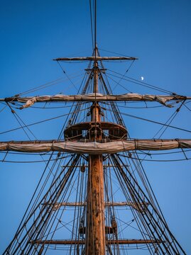 The mast and rigging of a historic sailing ship against a clear blue sky with a sliver of moon