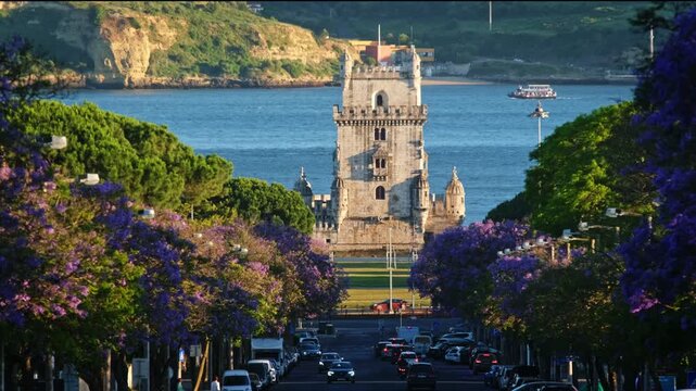 Jacaranda trees bloom with purple flowers near Belem Tower in the Belem district of Lisbon, Portugal. Evening sunlight warms the tower and the tree-lined avenue.