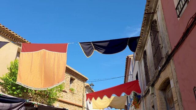 Low angle view of a narrow street decorated with colorful fabrics and banners in the historic Spanish village of Lliber. Old stone houses, tile roofs, and bright blue clear sky during the traditional 