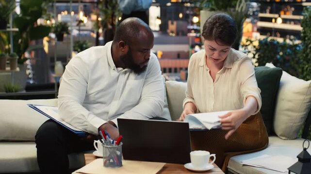 Close up of businesspeople reading a printed report during a rooftop lounge meeting, while colleagues analyze paperwork files and compare project data on an urban rooftop facing city offices.