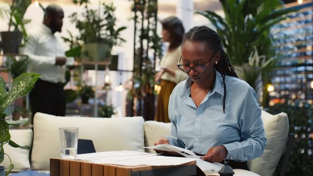 Businesswoman reviews printed reports from an outdoor workspace sofa surrounded by greenery, comparing project notes on a rooftop patio above downtown.