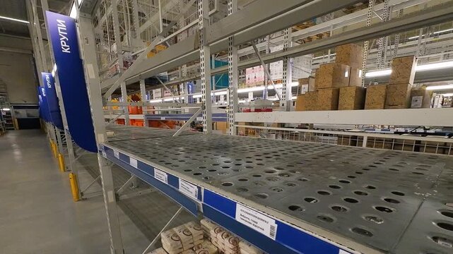 Empty metal shelves inside a large wholesale supermarket warehouse storage area show perforated storage racks cleared out due to supply chain disruptions, reflecting a product deficit.