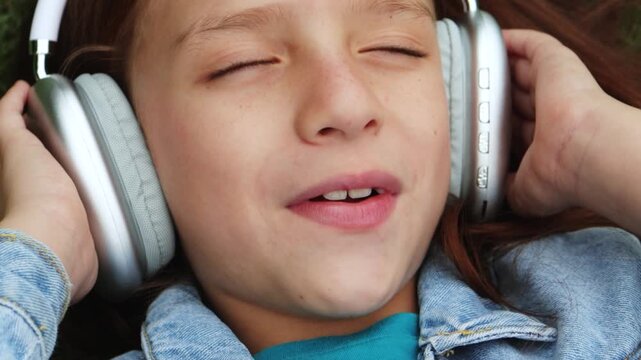 Child girl singing and listening to playlist music with headphones lying on grass - Technology trends concept