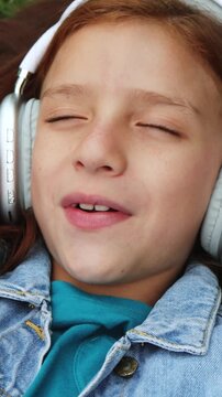 Child girl singing and listening to playlist music with headphones lying on grass - Technology trends concept - Vertical video