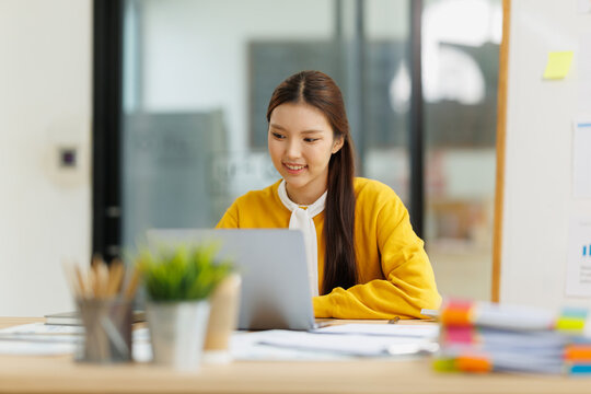 Young asian businesswoman smiling, enjoying successful remote work on laptop, achieving professional goals and productivity in a modern office environment