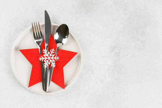 Christmas table setting with white plate and cutlery arranged in red star shaped cutlery holder decorated with snowflake on light gray textured background with copy space, elegant holiday backdrop.