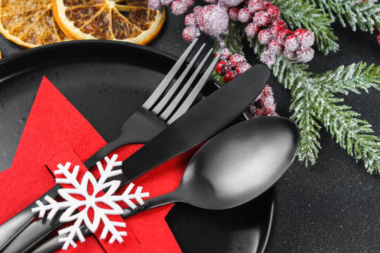 Holiday table setting with black cutlery arranged in red star shaped cutlery holder decorated with white snowflake, showy Christmas tree branch on dark textured background