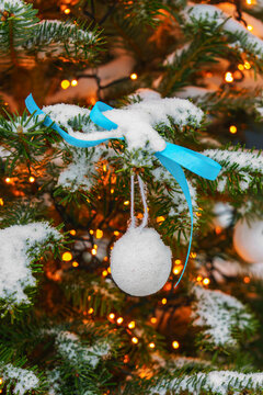 Snow covered Christmas tree branches decorated with white ornaments and a blue ribbon on glowing fairy lights colorful bokeh background. Cozy winter holiday atmosphere.